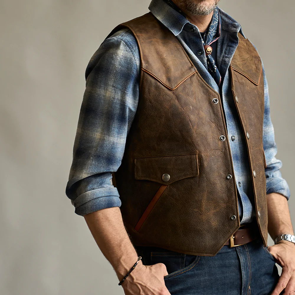 Man wearing a brown leather vest over a blue plaid shirt with a neutral background