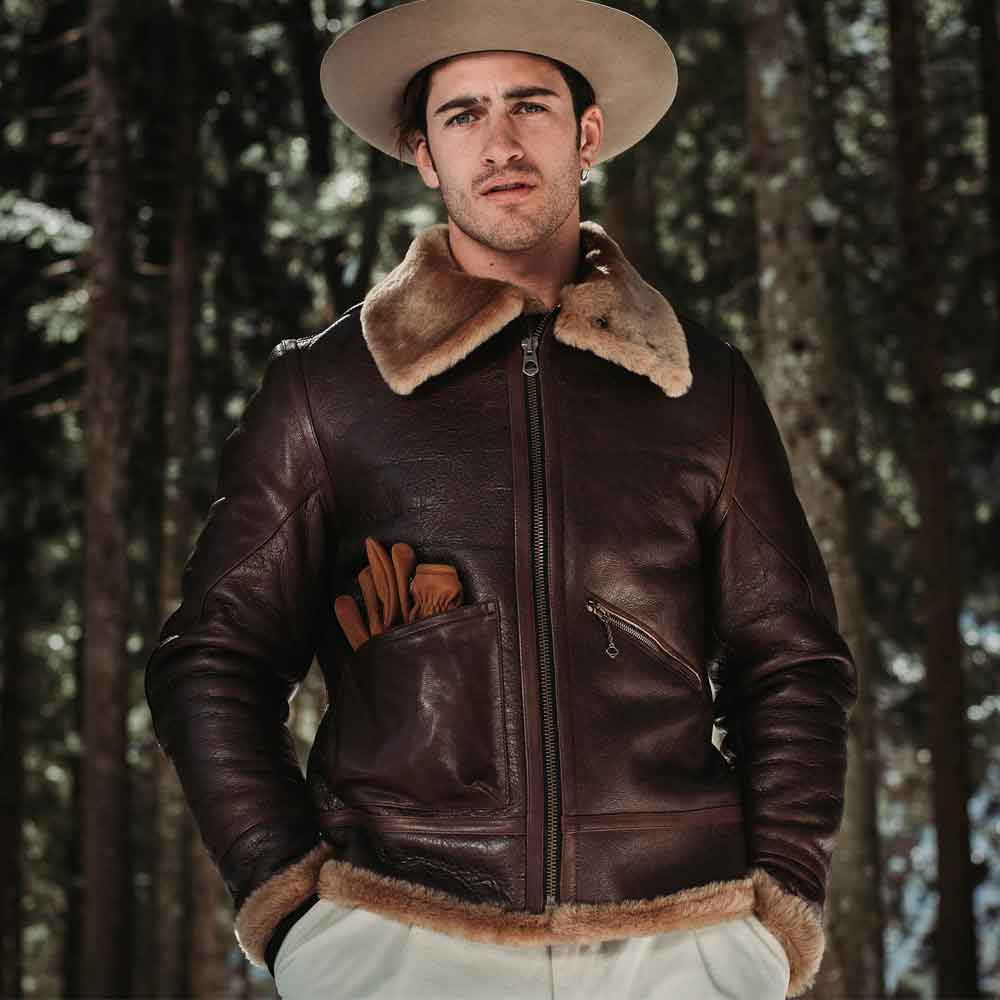 Man wearing a brown leather jacket with fur trim and a hat in a forest setting