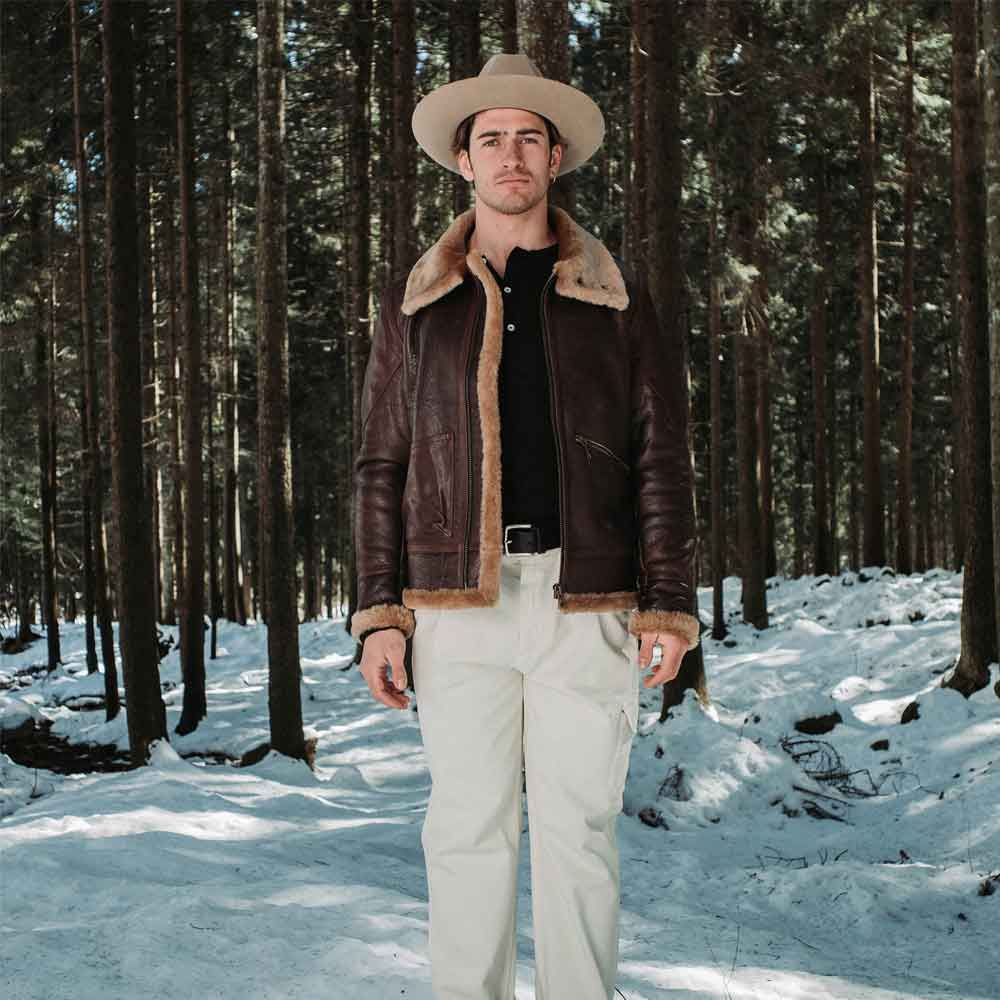 Man wearing a brown leather jacket with fur lining, white pants, and a hat standing in a snowy forest.