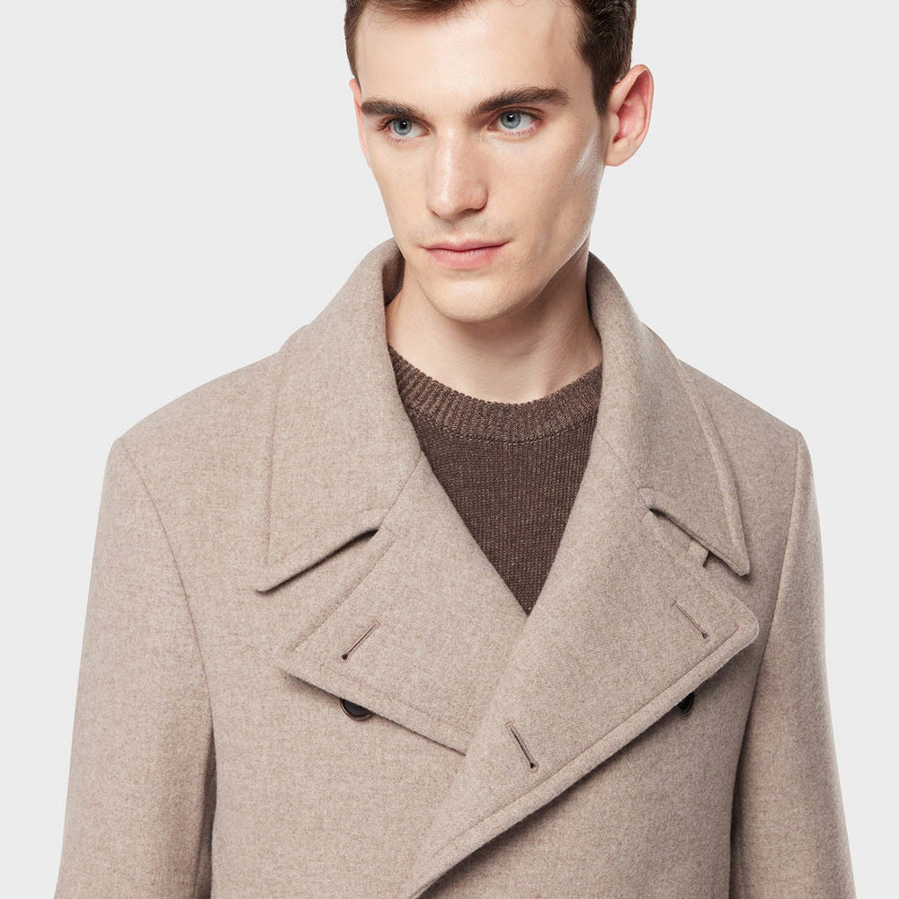 Man wearing a beige peacoat over a brown sweater on a light gray background