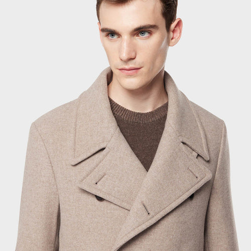 Man wearing a beige peacoat over a brown sweater on a light gray background