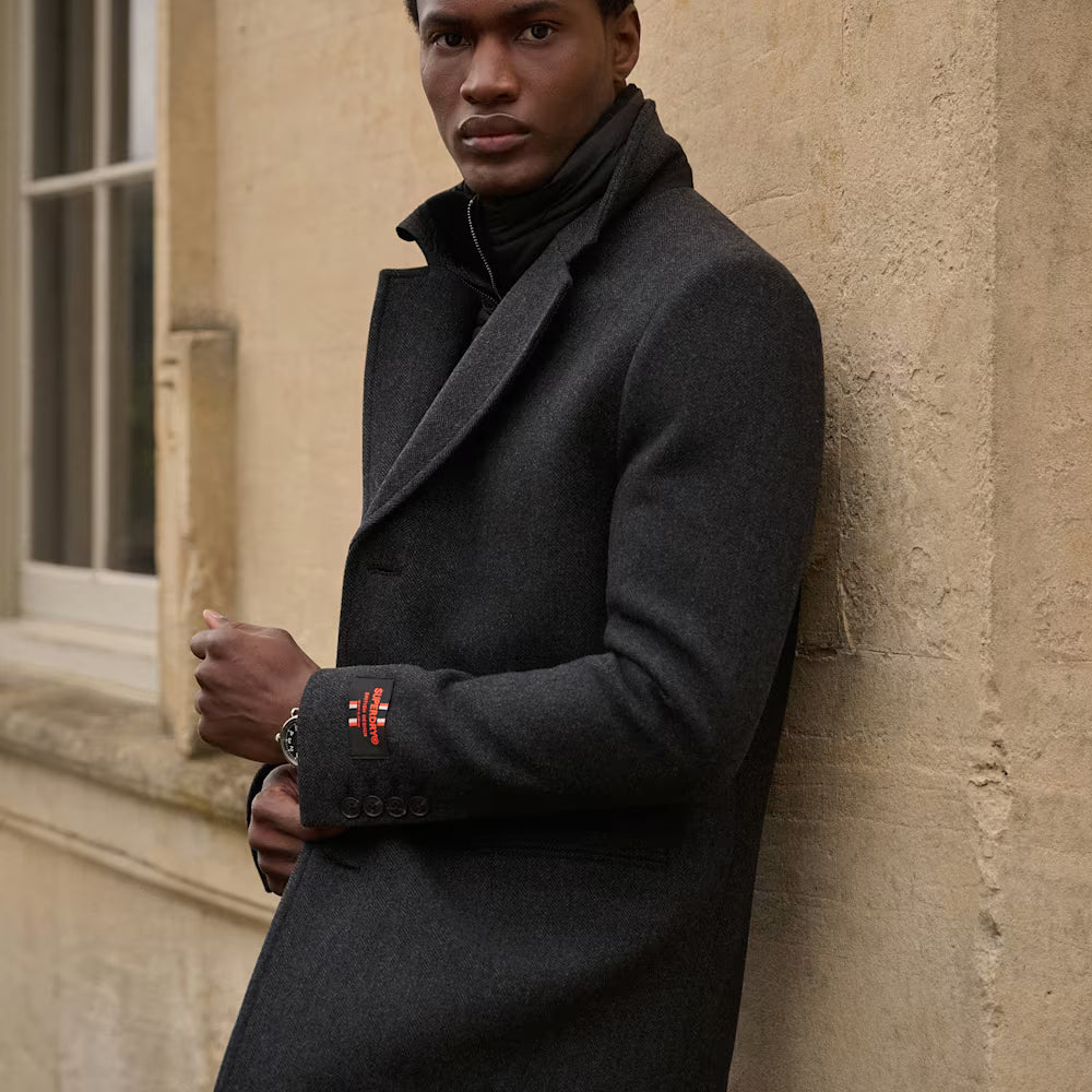 Man wearing a dark peacoat, leaning against a beige wall.