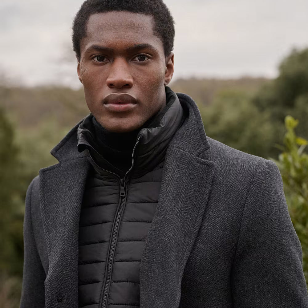 Man wearing a black peacoat over a black puffer jacket with a blurred natural background