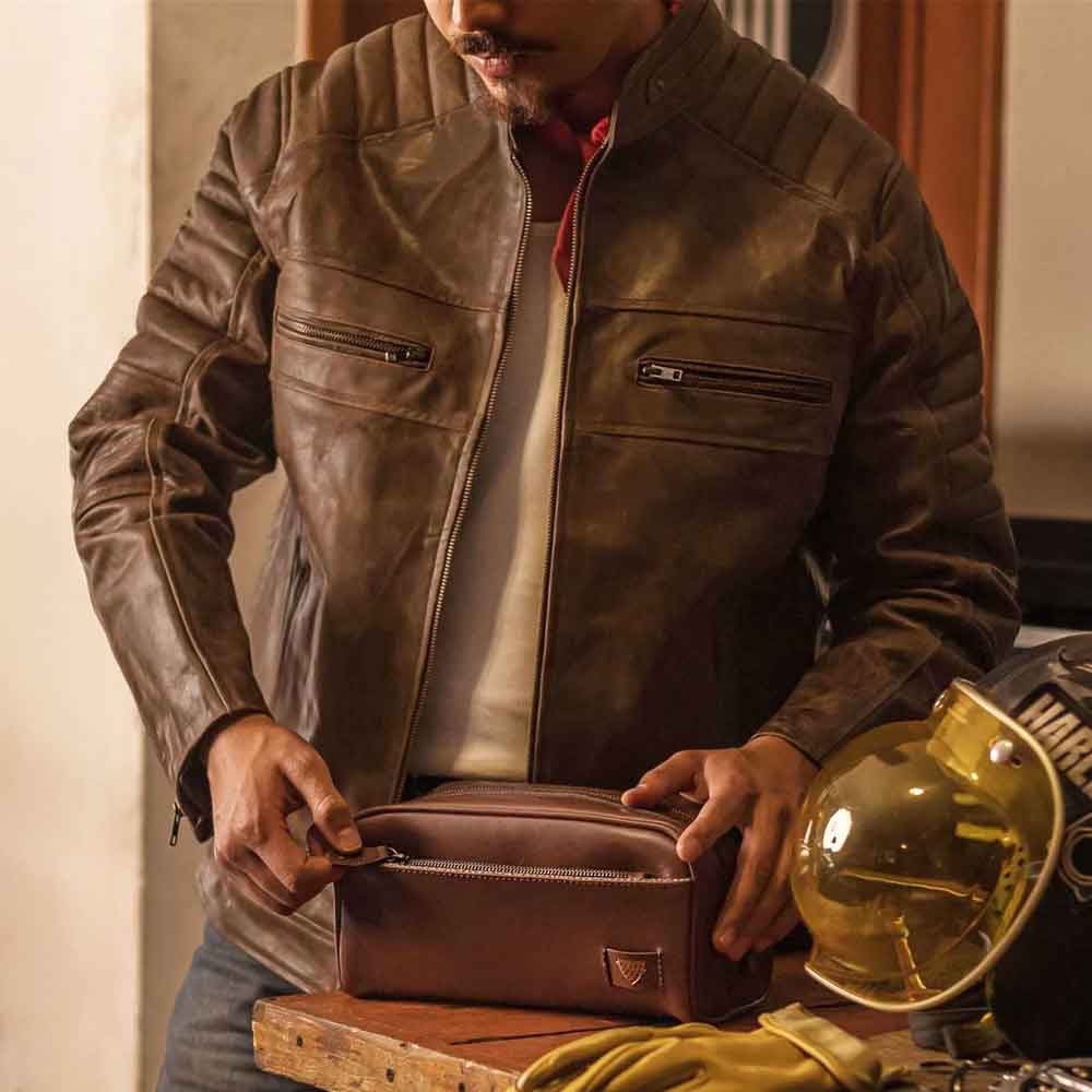 Man in a brown leather jacket holding a brown leather pouch with a helmet in the background.