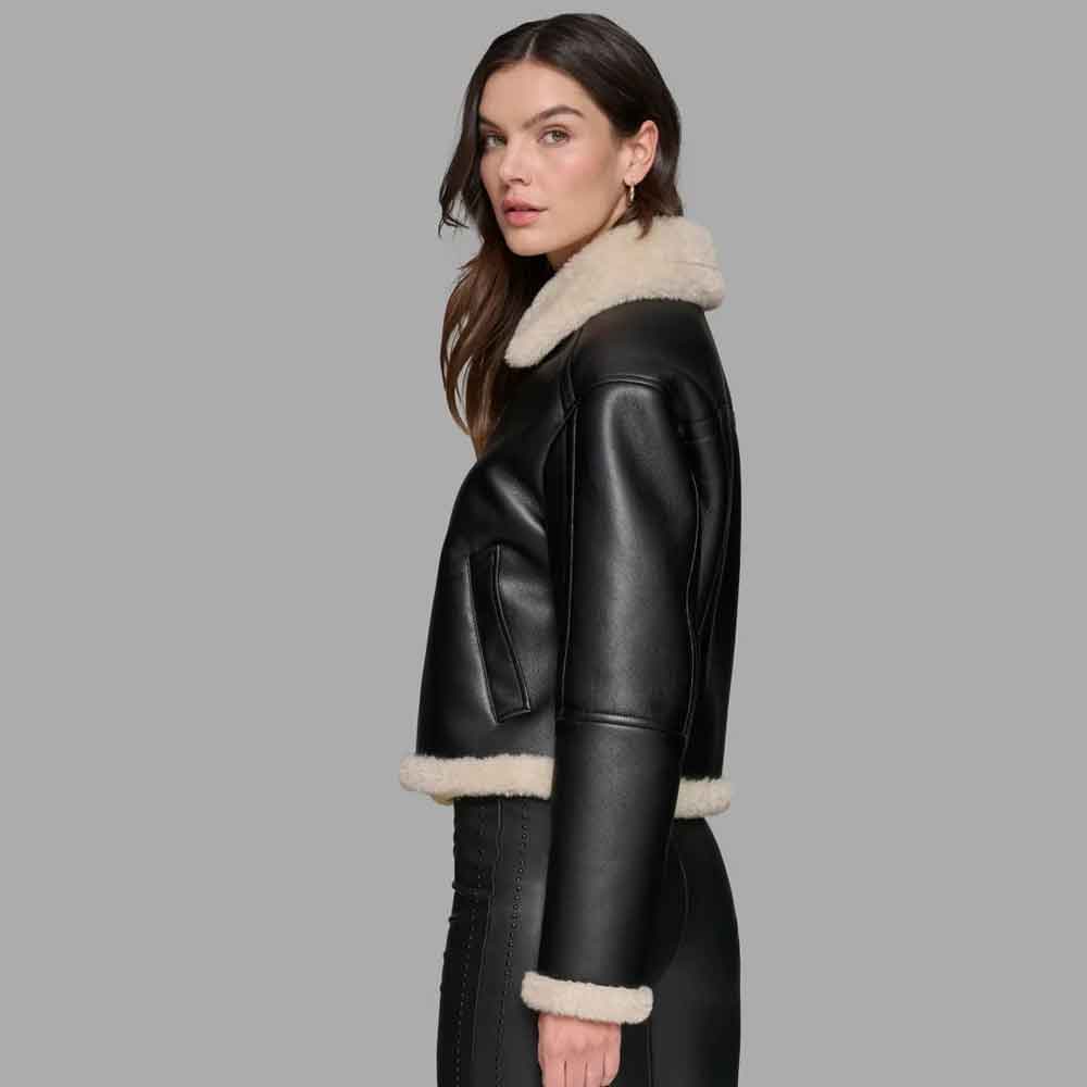 A woman modeling a black leather aviator jacket with a thick cream shearling-style collar, hem, and cuffs, shown from a side profile against a solid gray background.