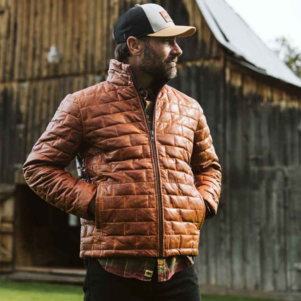 Man wearing a brown quilted jacket and cap standing in front of a rustic wooden building.