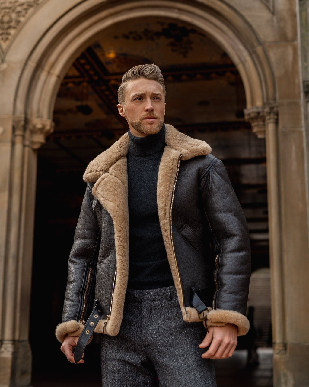 Man wearing a leather jacket with fur lining standing in an archway.