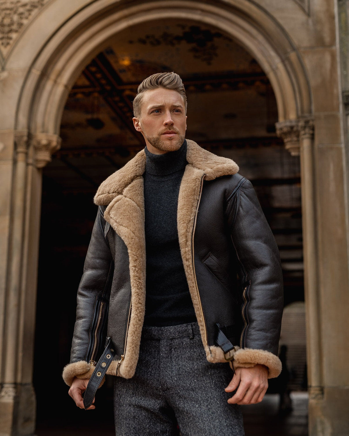 Man wearing a leather jacket with fur lining standing in an archway.